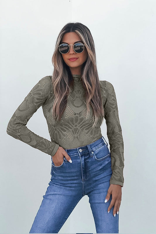 Woman in sunglasses wears a Green Mock Neck Fishnet Top and blue high-waisted jeans, standing against a white background.