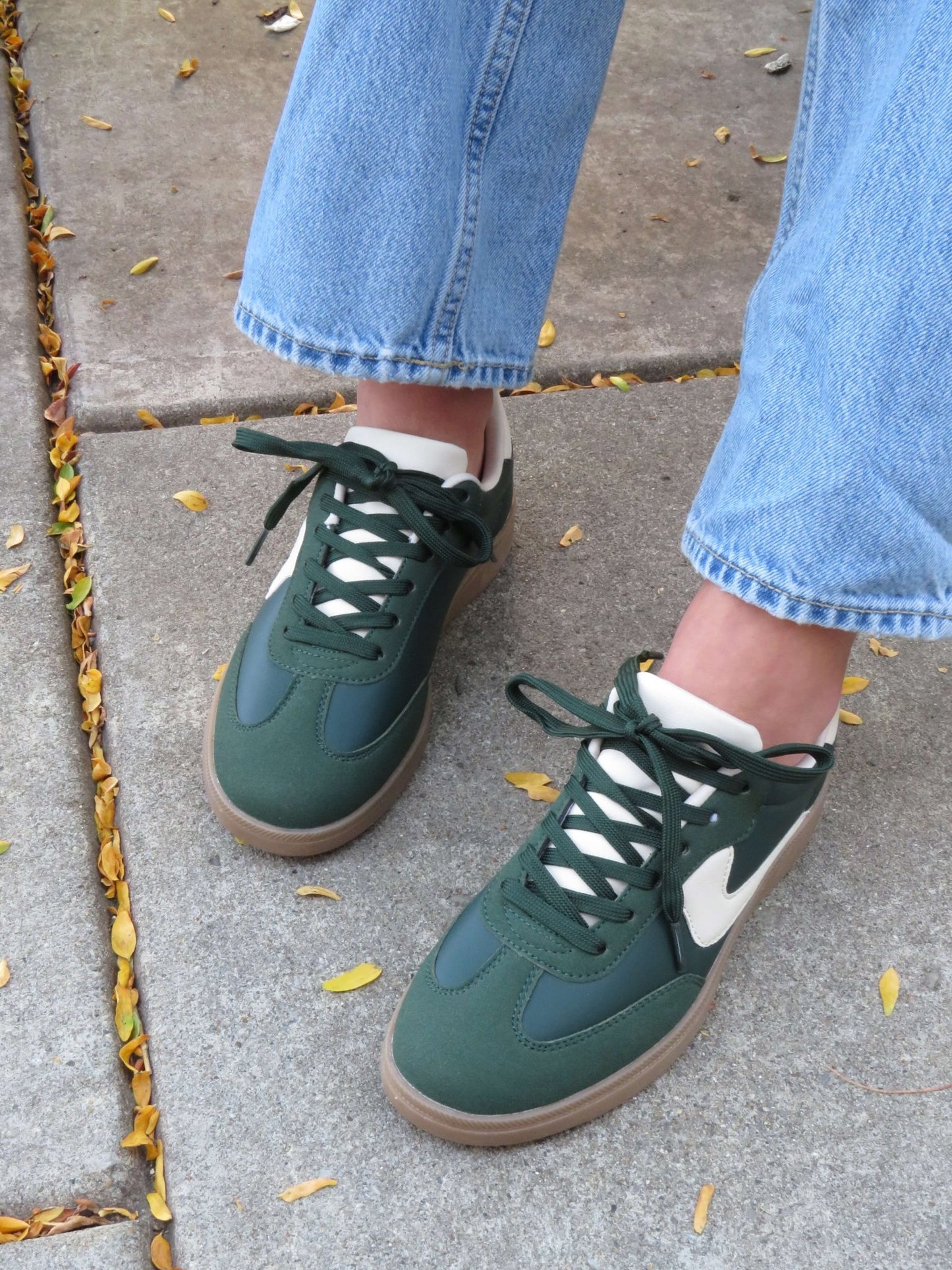 Person in light blue jeans and Green Remus Lace Up Sneakers - DOORBUSTER, standing on a sidewalk with scattered yellow leaves.