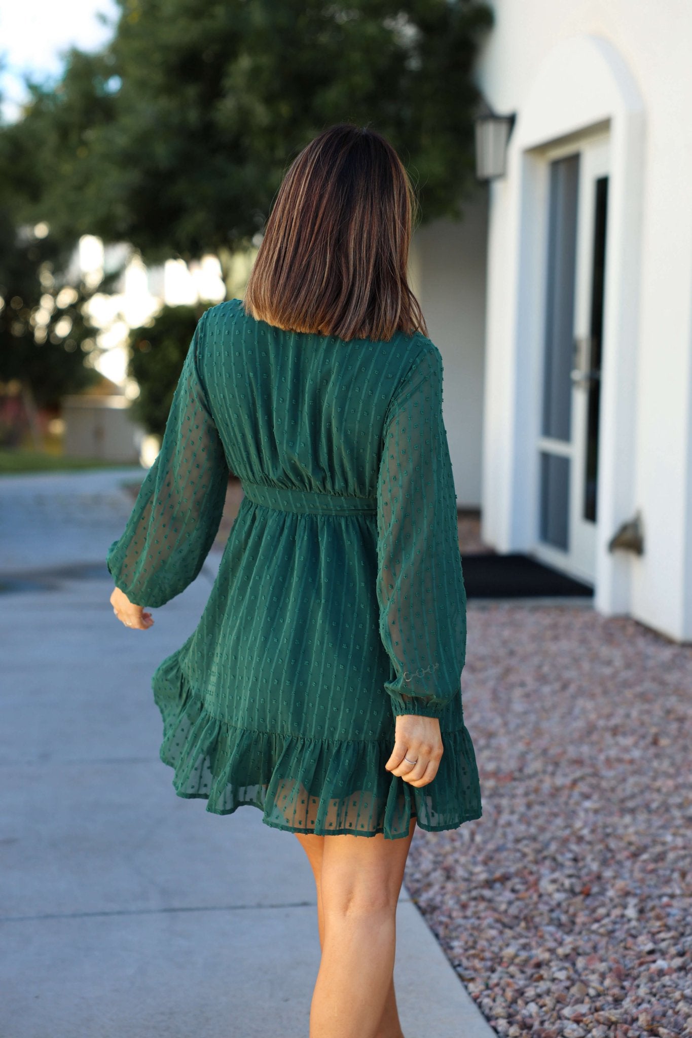 A woman walks on a sidewalk, her back to the camera, wearing a trendy Green Swiss Dot Ruffled Mini Dress.