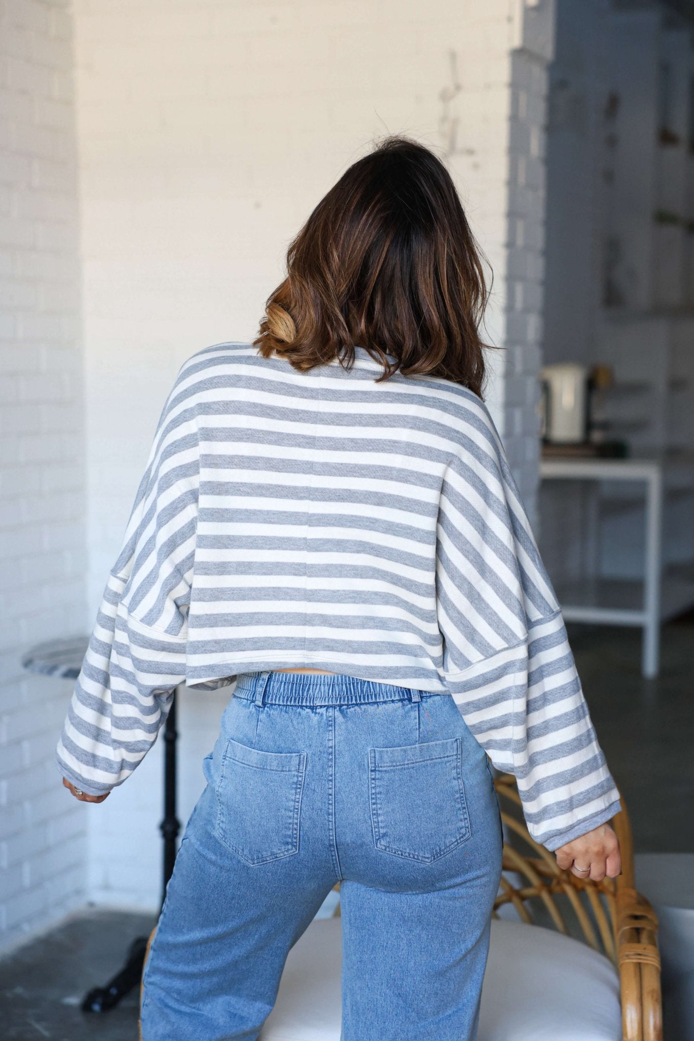 A person with shoulder-length brown hair wears a Grey and White Striped Boxy Pullover and blue jeans, back to camera indoors.
