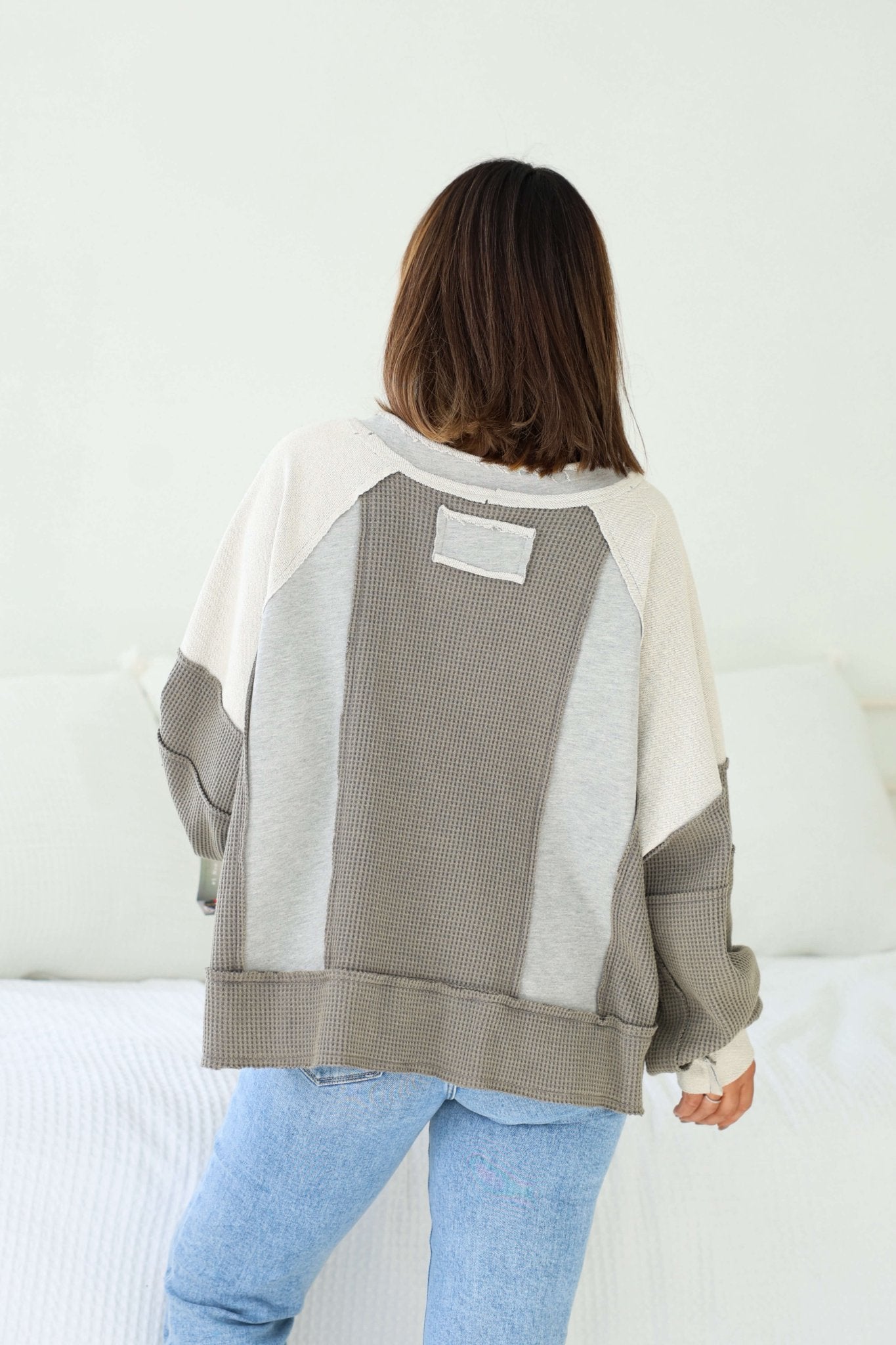 A person with shoulder-length hair wears the Grey Contrast Frayed Edge Pullover and blue jeans in a bright, minimal room.