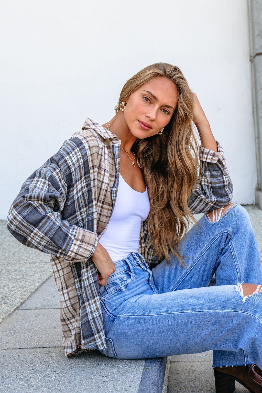Woman with long wavy hair wears the Grey Plaid Button Down Top over a white tank and ripped jeans while sitting outdoors.