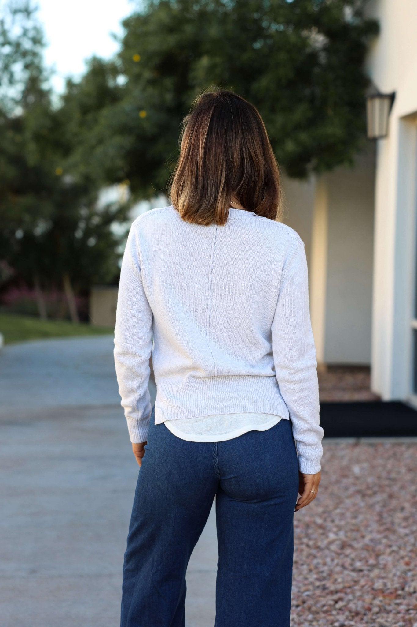 A woman with shoulder-length brown hair, wearing a grey V neck ribbed sweater and blue pants, stands outside facing away.
