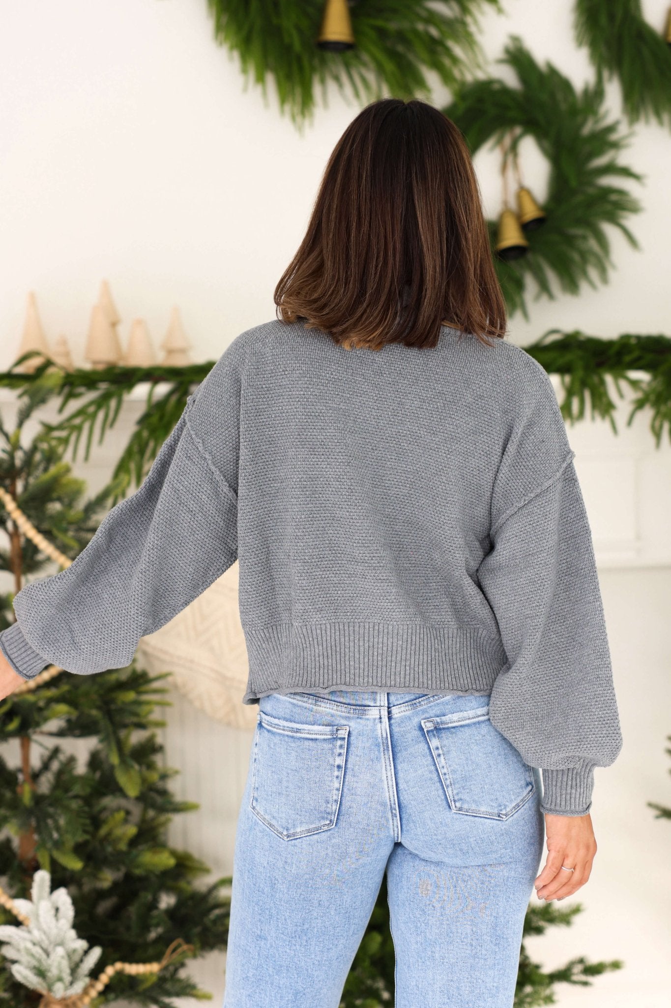 A woman in a Grey Waffle Knit Open Front Cardigan and light blue jeans stands with her back to the camera, among festive greenery.