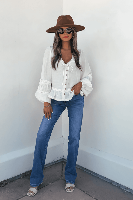 Woman in hat, sunglasses, Hadley Cream Ruffle Lace Trim Blouse, blue jeans, and sandals poses by a white corner wall.