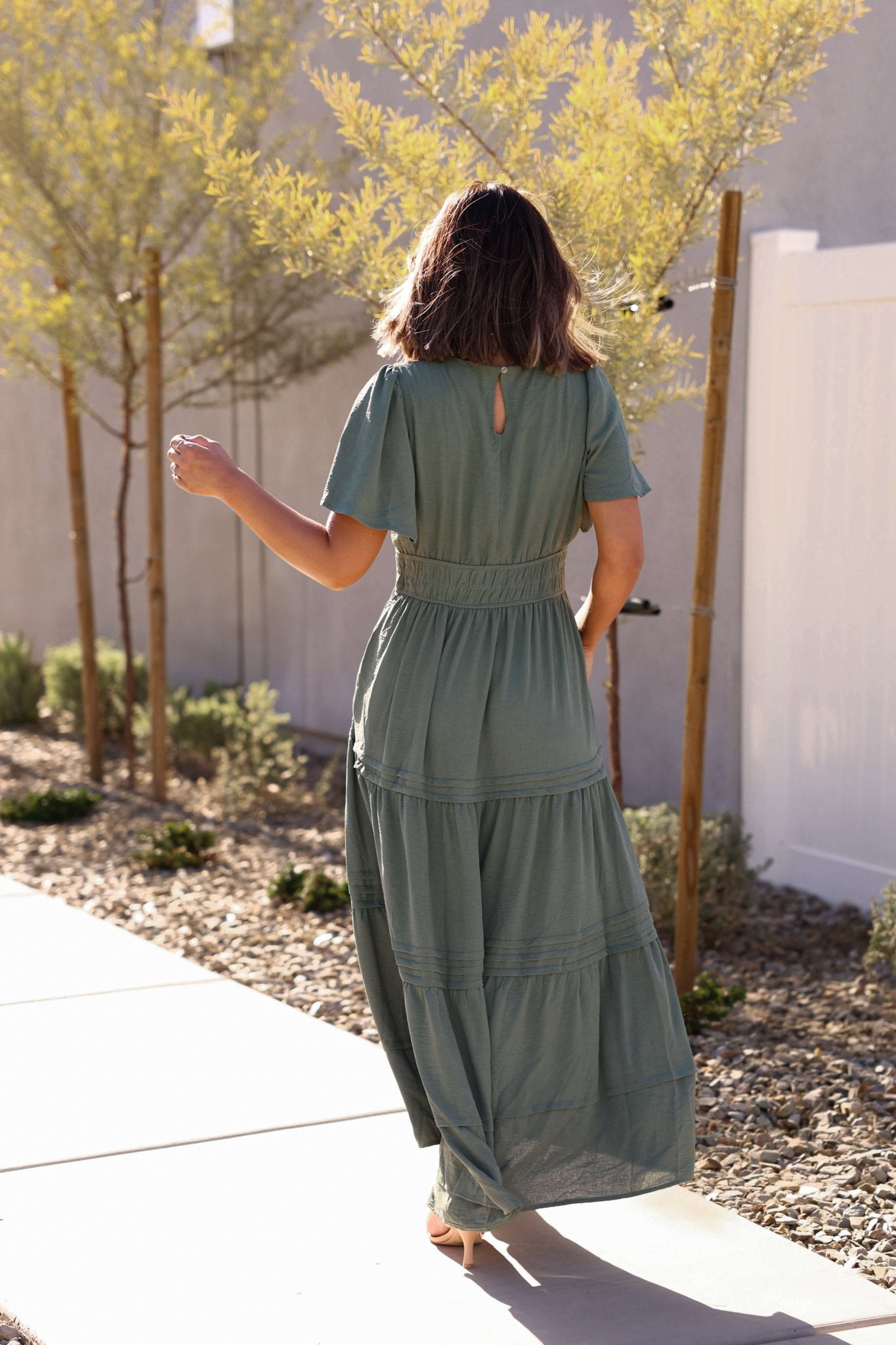 Woman in the Hannah Surplice Tiered Maxi Dress - Olive walks on a sunlit sidewalk surrounded by trees and plants.