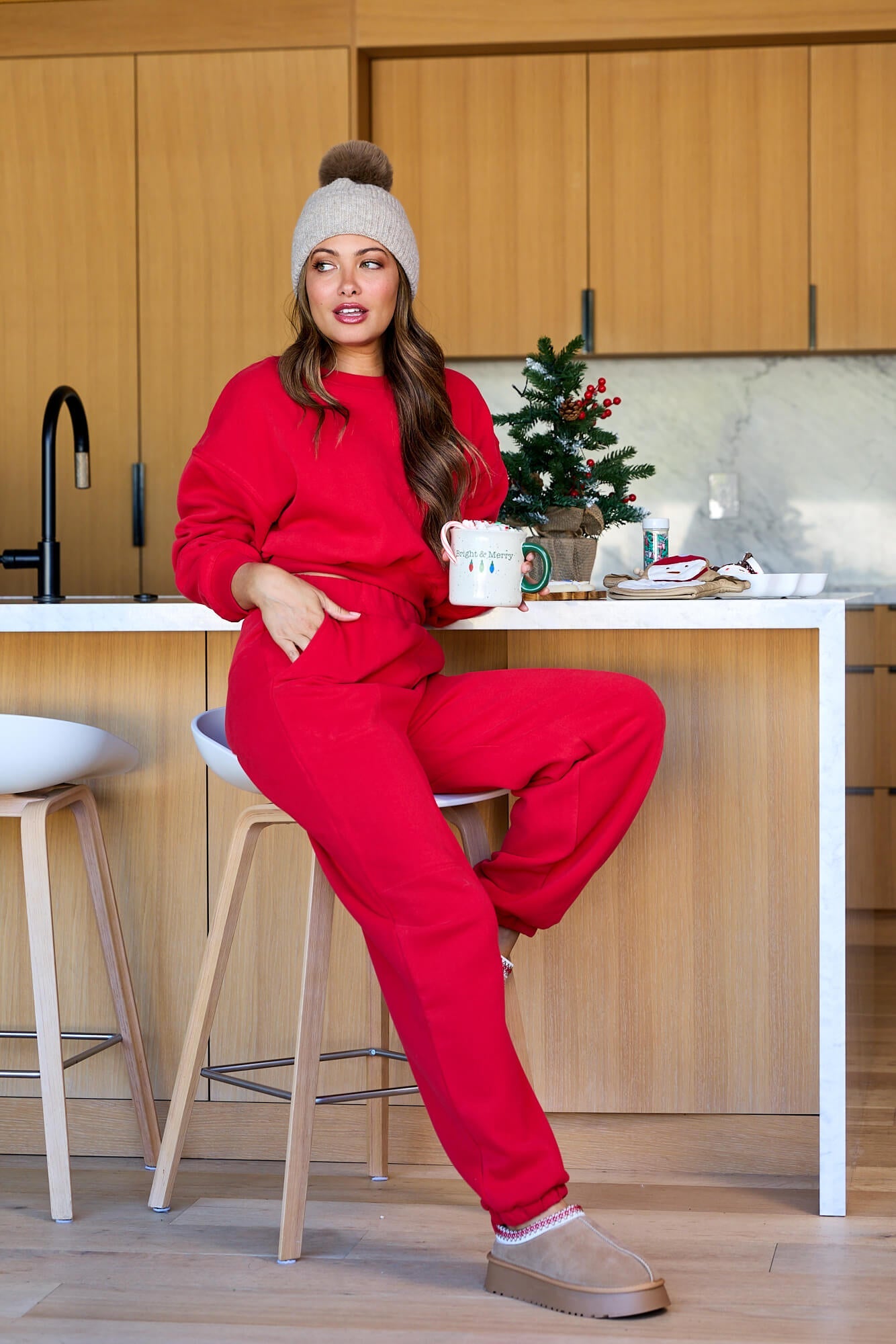 Woman in Holiday Red Lounge Jogger Sweatpants - FINAL SALE sits on a kitchen stool, holding a mug by a decorated Christmas tree.