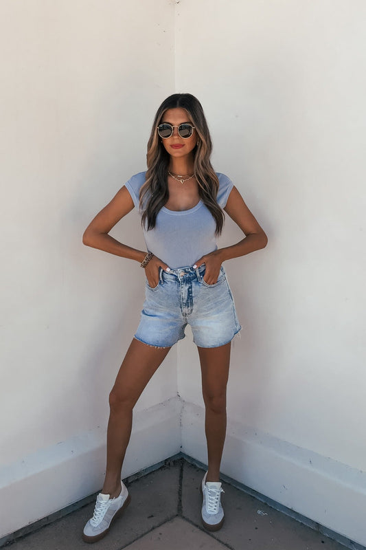 Woman wears an Ice Blue Scoop Neck Ribbed Top with denim shorts and white sneakers, standing by a white wall, hands on hips.