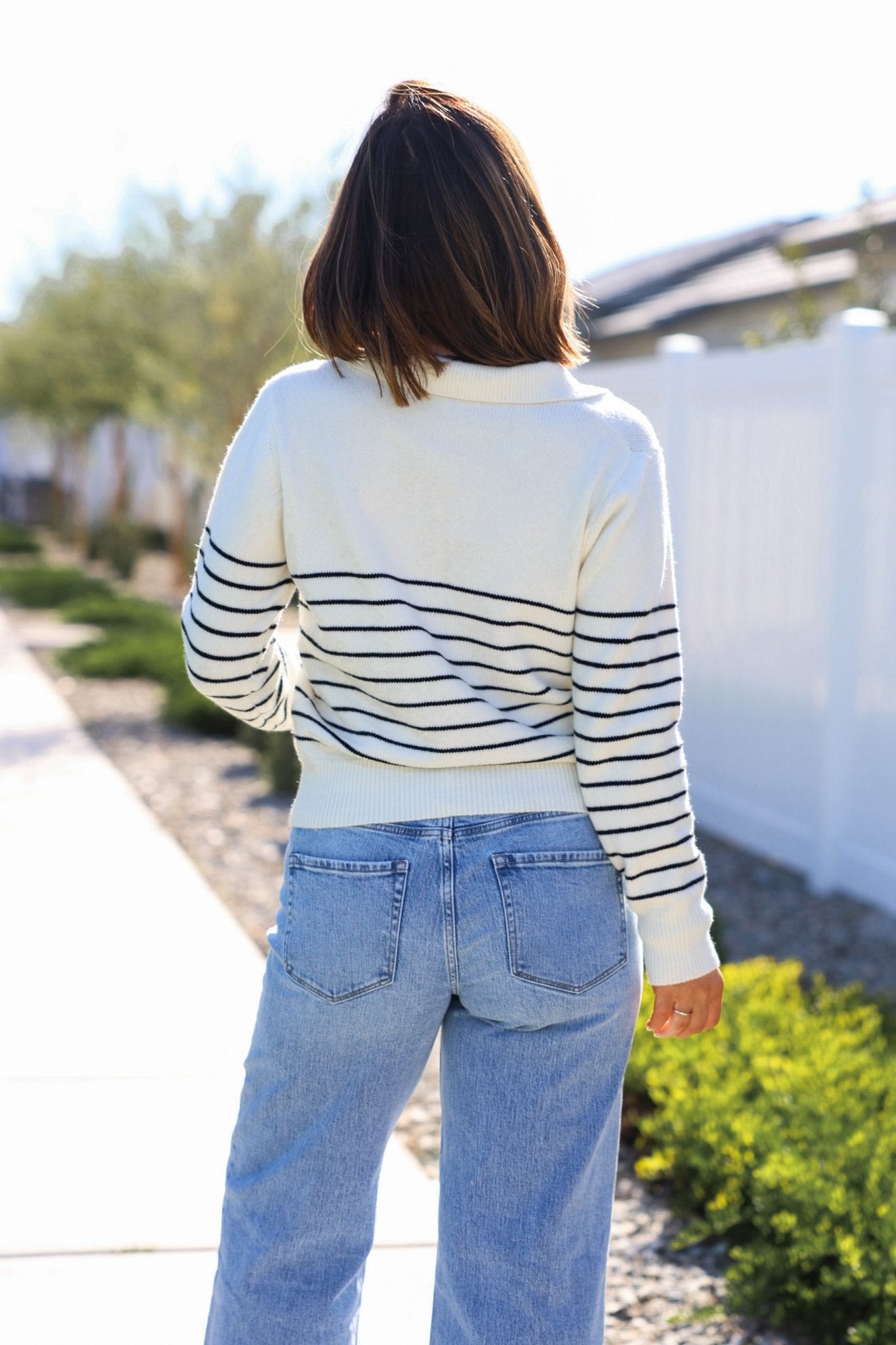 A woman with shoulder-length brown hair wears the Ivory and Black Stripe Ribbed Sweater - FINAL SALE and blue jeans on a sidewalk.
