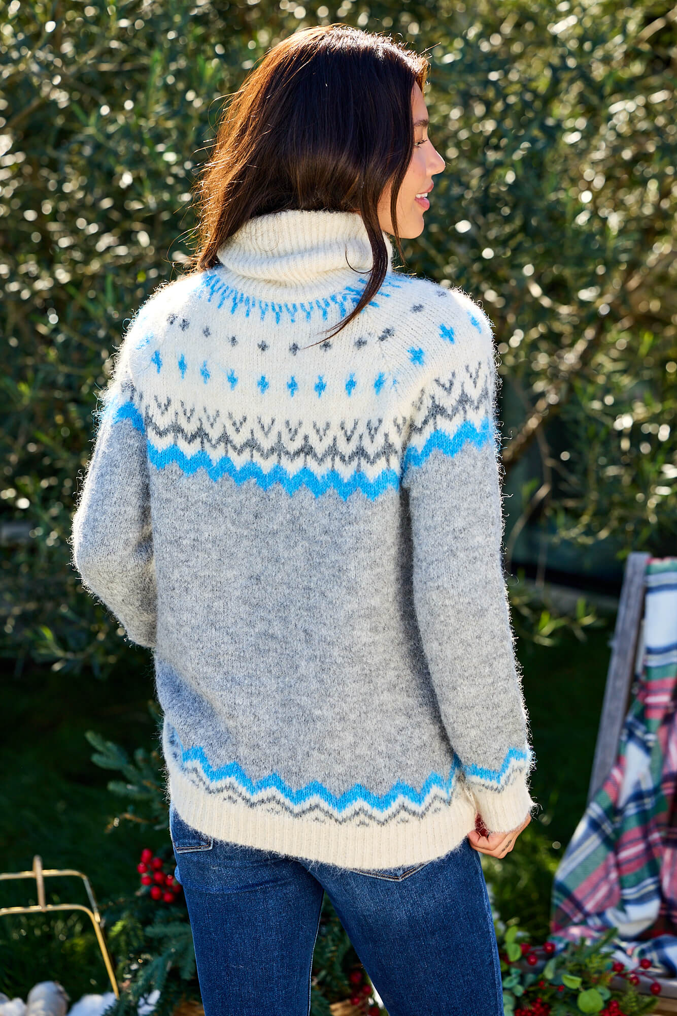 A woman stands outdoors amid greenery in the Ivory and Grey Fair Isle Turtleneck Sweater-FINAL SALE, facing away.