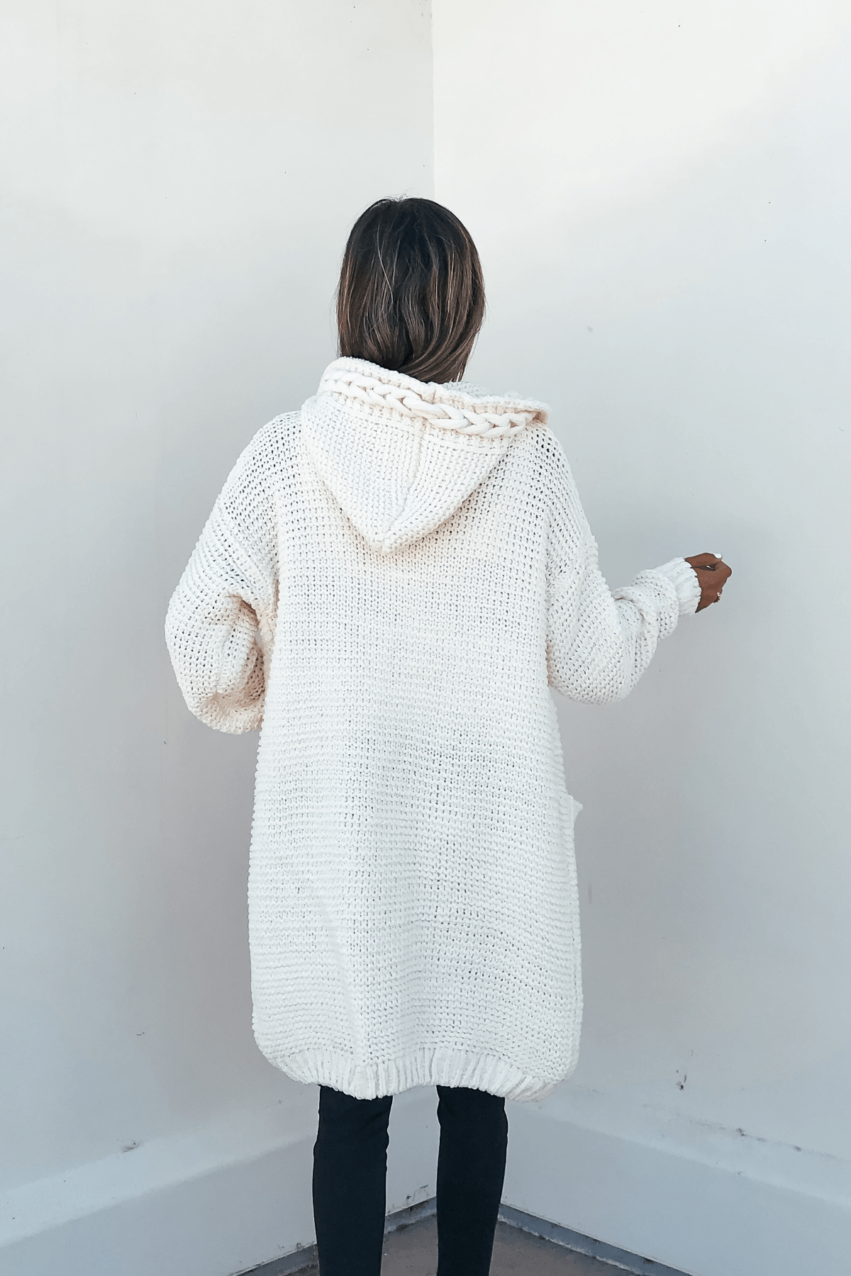 Person faces away from the camera in an Ivory Braided Trim Hooded Cardigan and black pants against a white wall.
