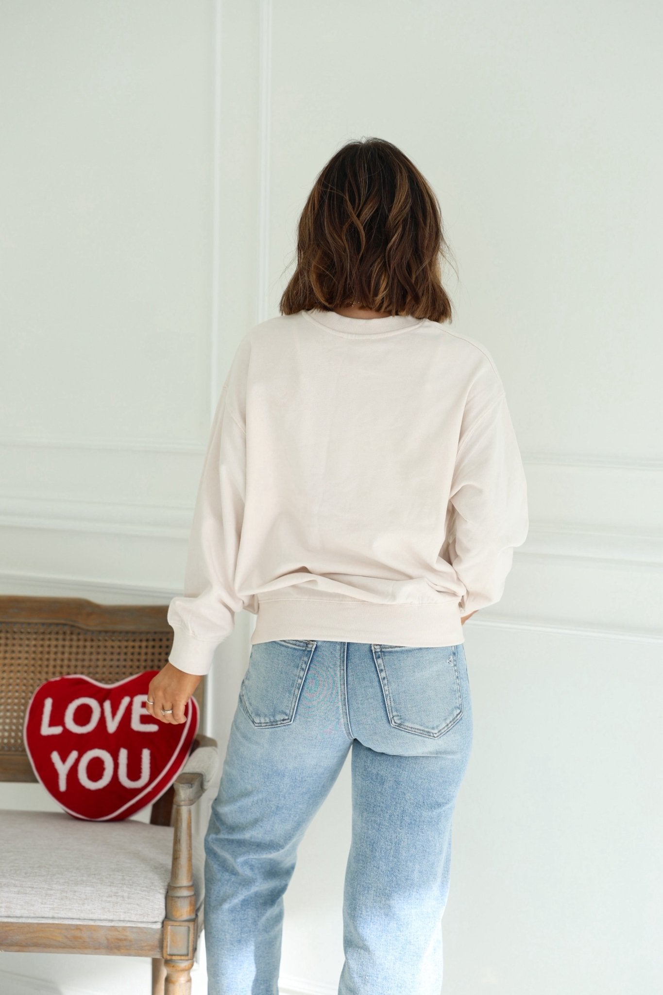 A woman in a Light Beige Cupid Cowboy Graphic Sweatshirt and jeans stands by a chair with a red heart-shaped LOVE YOU pillow.