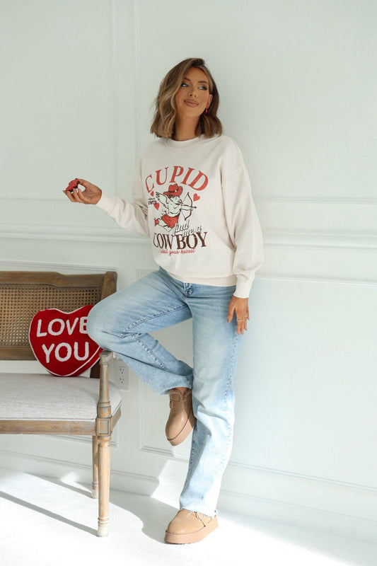 A woman in a Light Beige Cupid Cowboy Graphic Sweatshirt and jeans stands by a bench with a red LOVE YOU pillow, holding a small object.