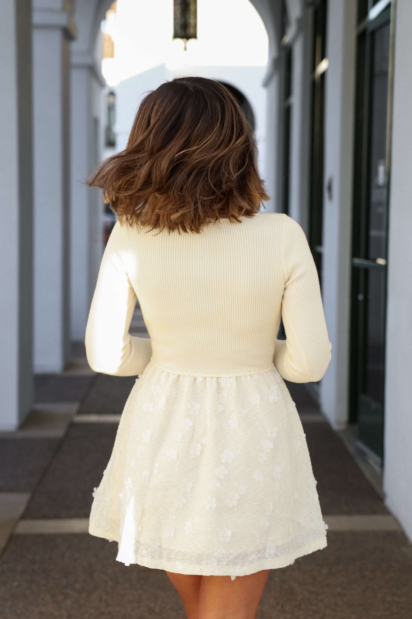 A woman in an Ivory Floral Textured Mini Dress walks down a sunlit corridor, her shoulder-length brown hair glowing in the light.
