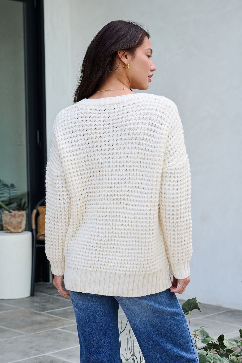 A woman with long brown hair models the Ivory Textured Cable Knit Sweater and blue jeans, shown from behind on a patio.