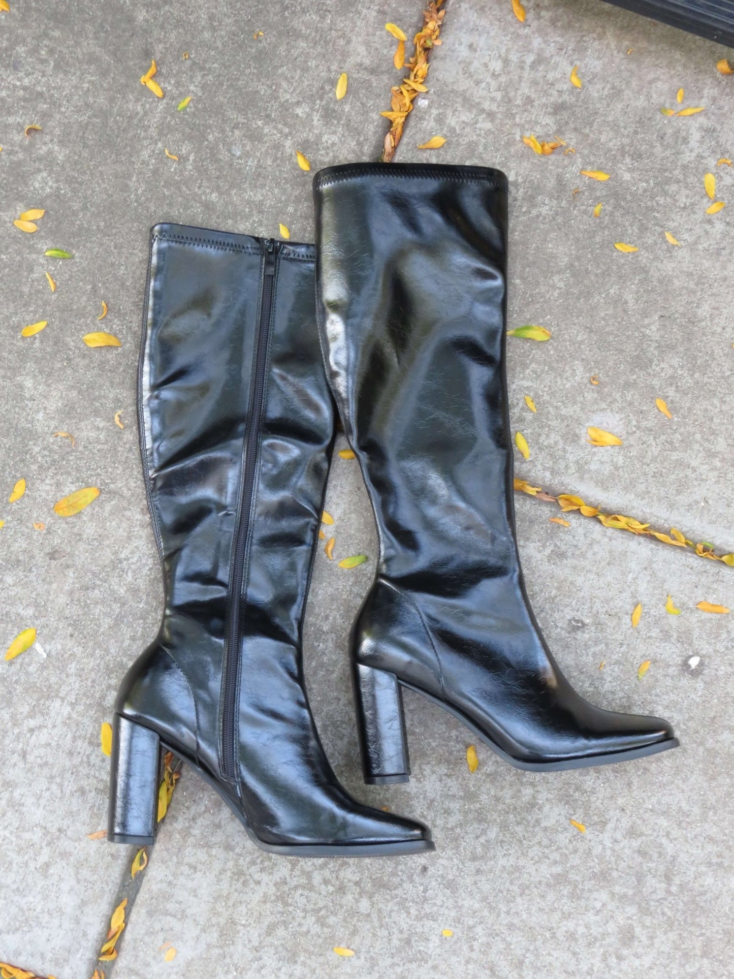 The Ivy Black Faux Leather Heeled Knee High Boots with zippers rest on concrete, surrounded by scattered yellow leaves.