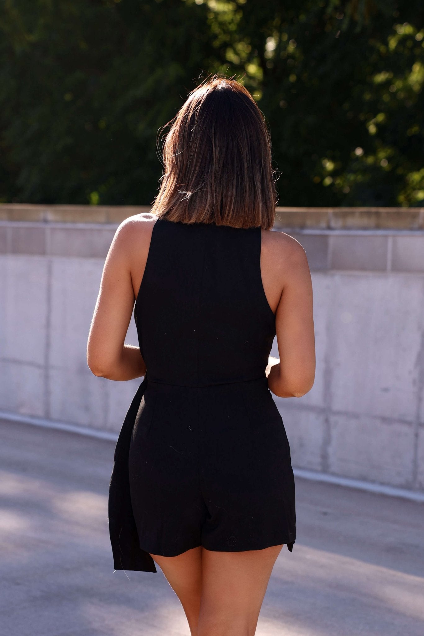 A woman in the Jameson Black Belted Romper with shoulder-length brown hair walks outdoors, facing away from the camera.