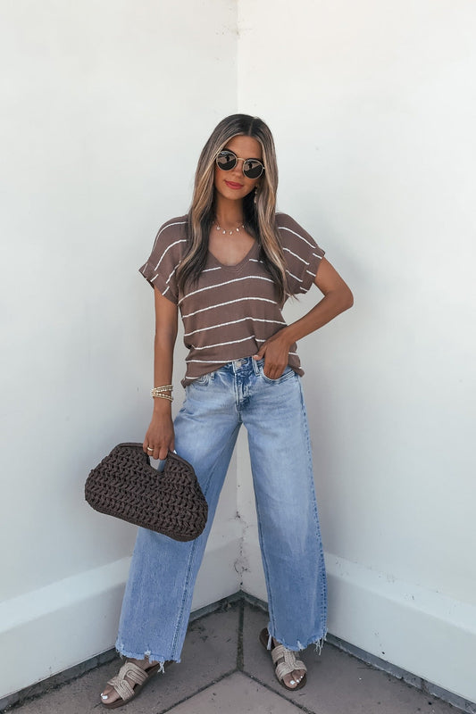 Woman in sunglasses wears the Jessica Striped Top - Mocha with wide-leg jeans, sandals, and a woven bag against a white wall.