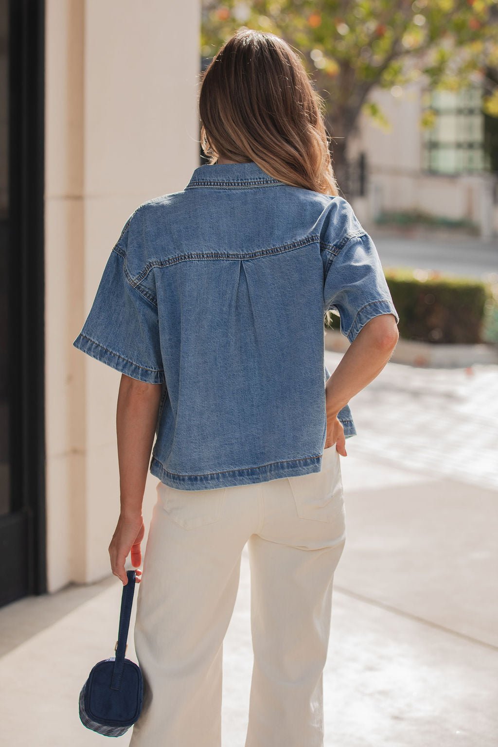 A woman with long hair wears the Jessie Denim Button Up Shirt and white pants, holding a small blue bag outdoors.