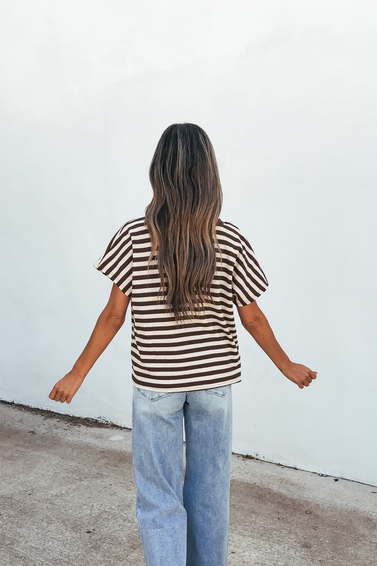 Woman with long wavy hair wears the Joan Striped Knit Top - Brown and jeans, standing outdoors facing a white wall, arms out.