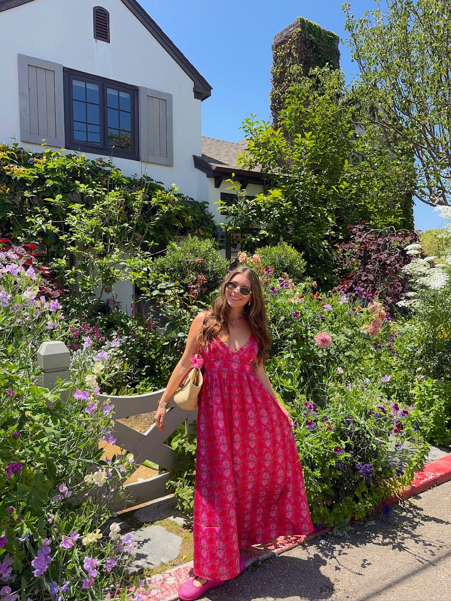 Woman wearing the Joyful Pink Floral Satin Maxi Dress stands smiling in front of a lush garden and white house on a sunny day.