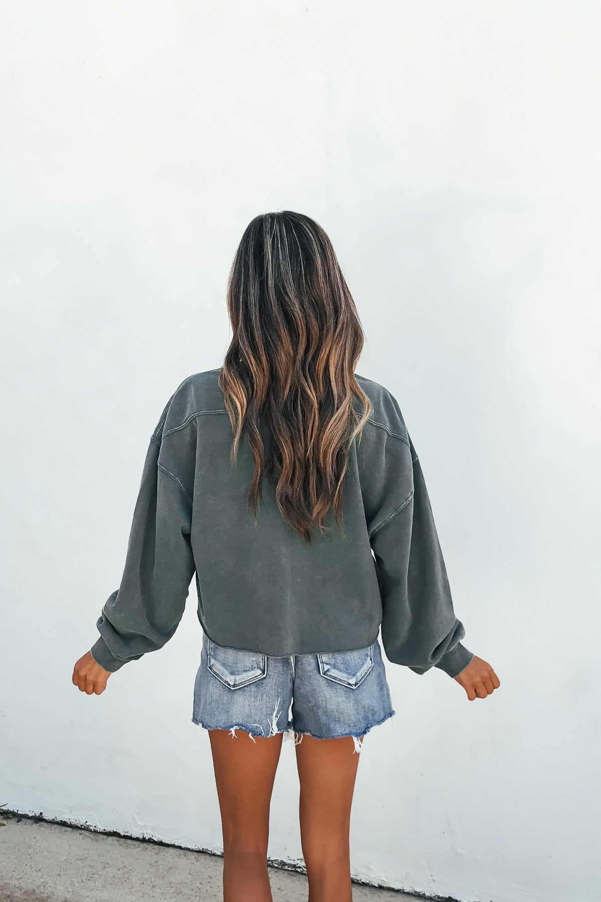 Person with long wavy hair wears a relaxed fit dark green Laid-Back Crewneck Pullover and denim shorts, back to camera, white wall.