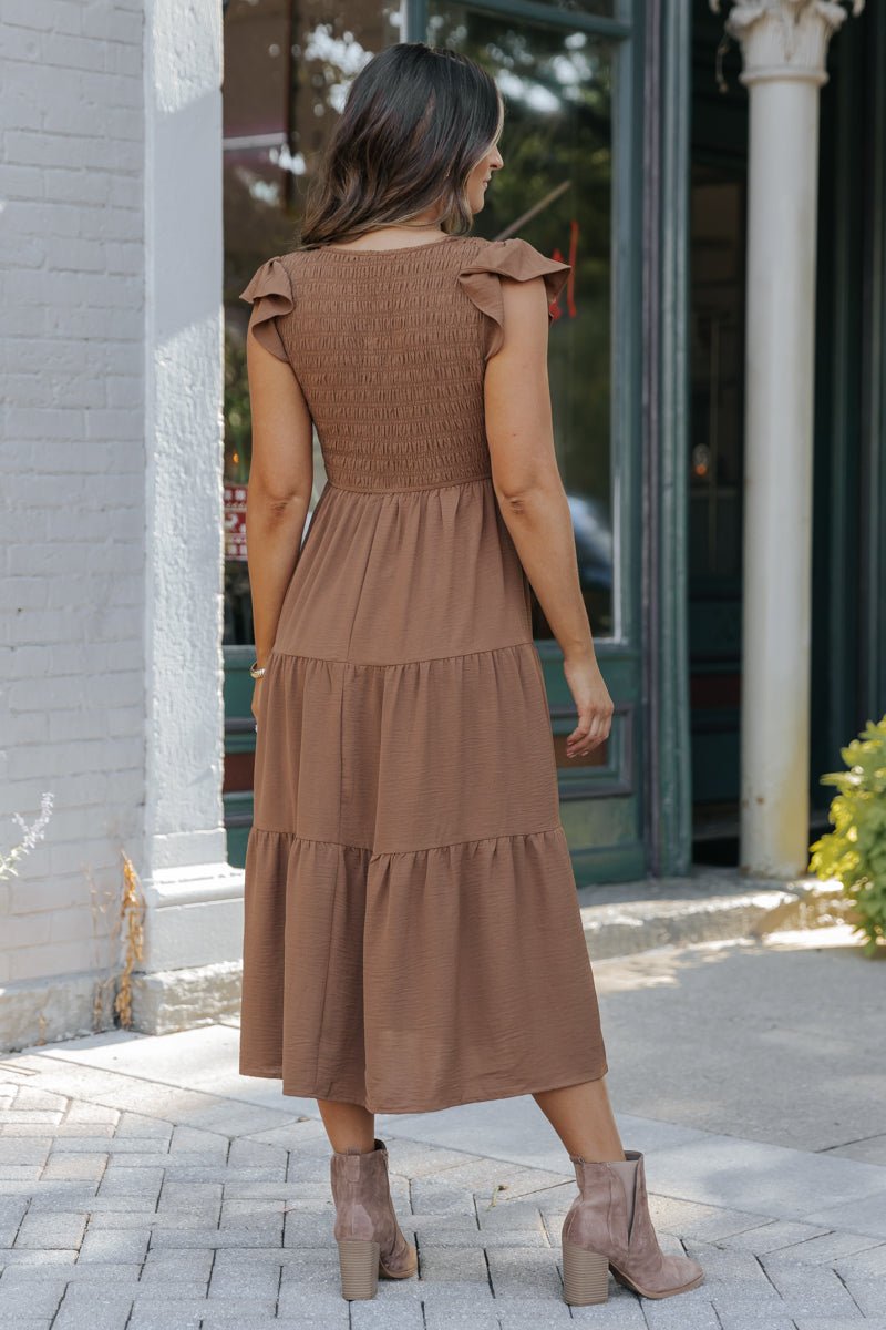 A woman in a Laney Brown Smocked Midi Dress - DOORBUSTER and taupe ankle boots stands outdoors, facing away.