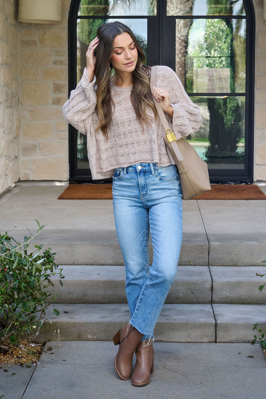 A woman in the Laurel Taupe Open Knit Sweater, light blue jeans, brown boots, and a tan bag stands on steps by a glass door.