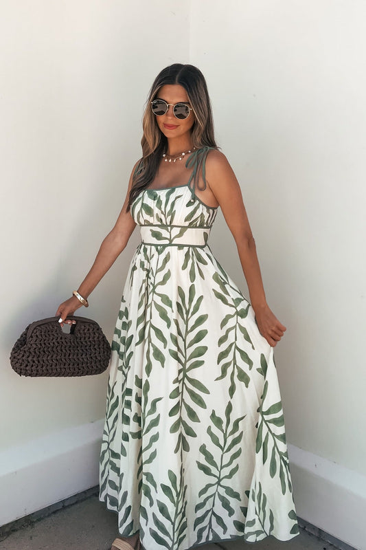 Woman in the Leaf Print Tie Strap Maxi Dress - Green, sunglasses on and woven bag in hand, stands against a white wall.