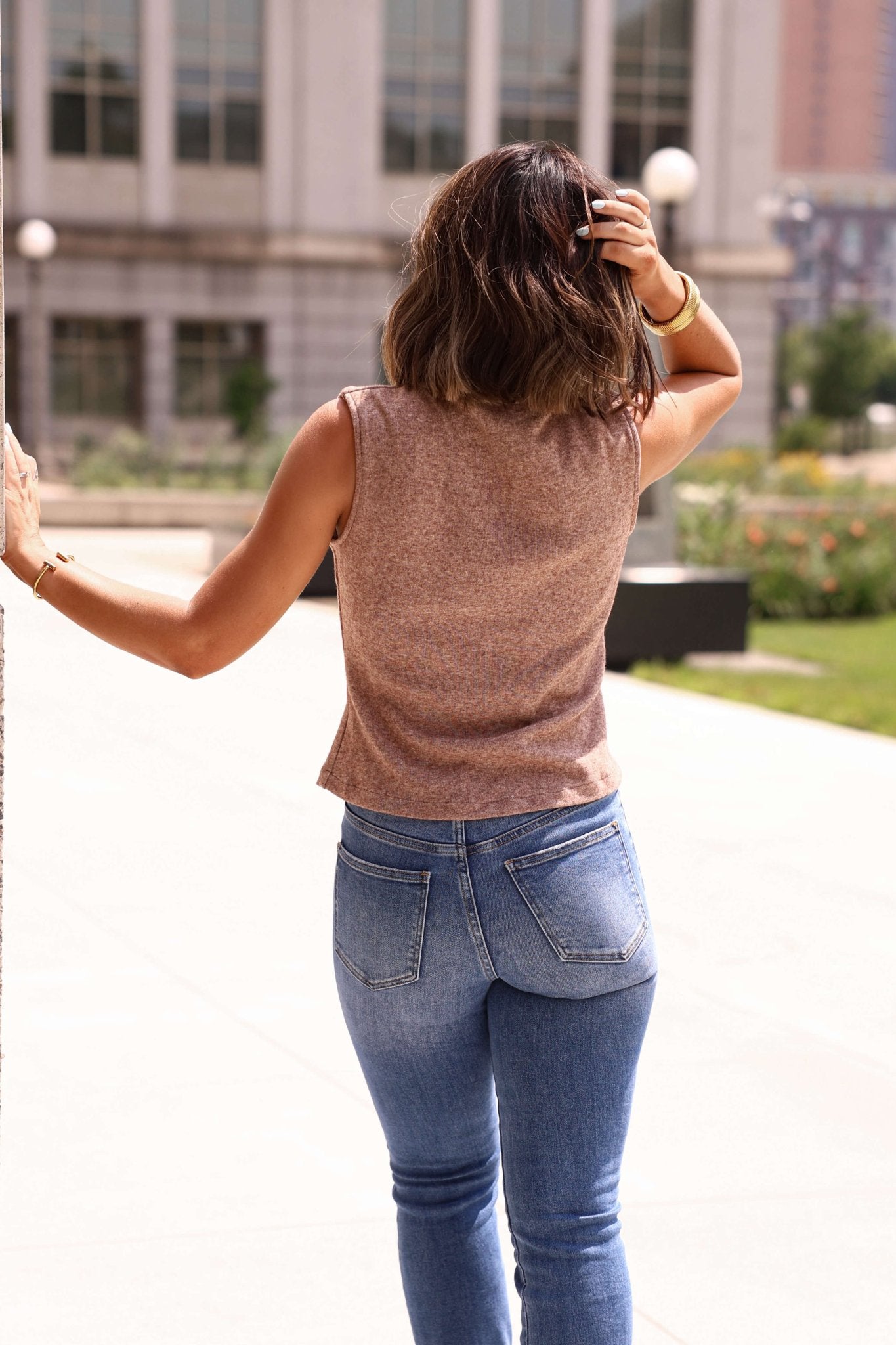 A woman in the Lenna Mocha Buttoned Vest - DOORBUSTER and jeans stands outdoors, facing away, hand in hair, one on a wall.