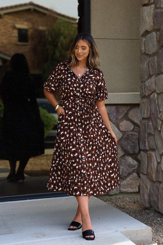 Woman in a Leopard Print Belted Midi Dress - Brown and black sandals stands by a stone wall, smiling with hand in pocket.