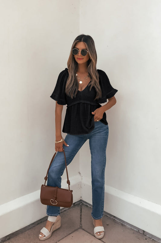 Woman in sunglasses, Lola Black Ruffled Babydoll Top, blue jeans, sandals, with a brown handbag by a white corner wall.