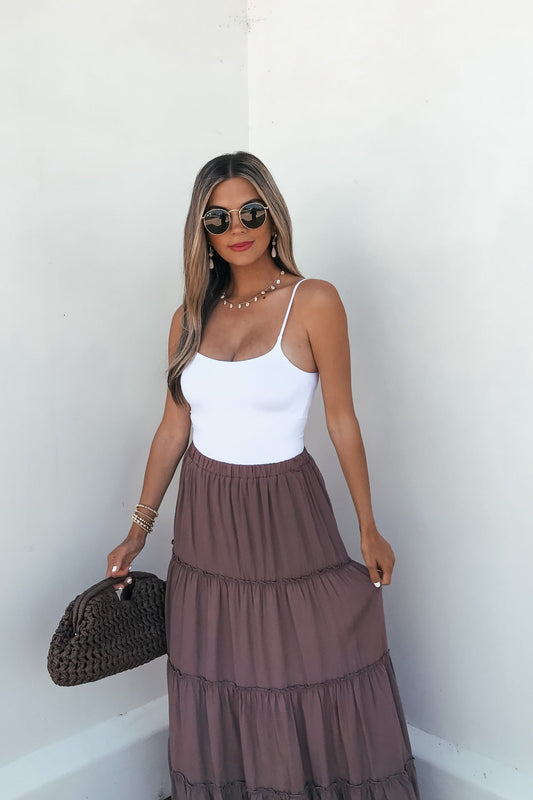 Woman in sunglasses, white tank, and Lori Ruffle Tiered Maxi Skirt - Mocha, holds a woven bag against a white wall.