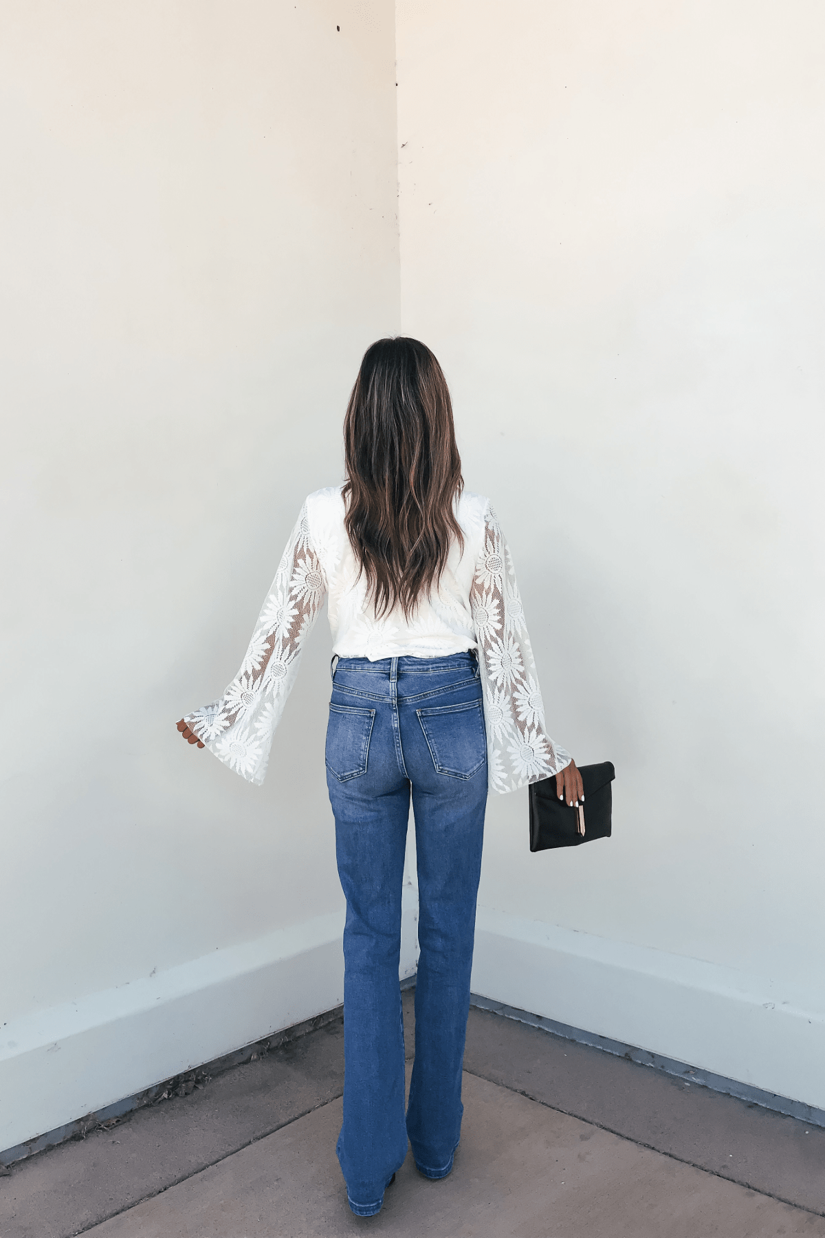 Woman facing a white wall, wearing flared jeans, the Louisa White Floral Lace Bodysuit, and holding a black clutch.