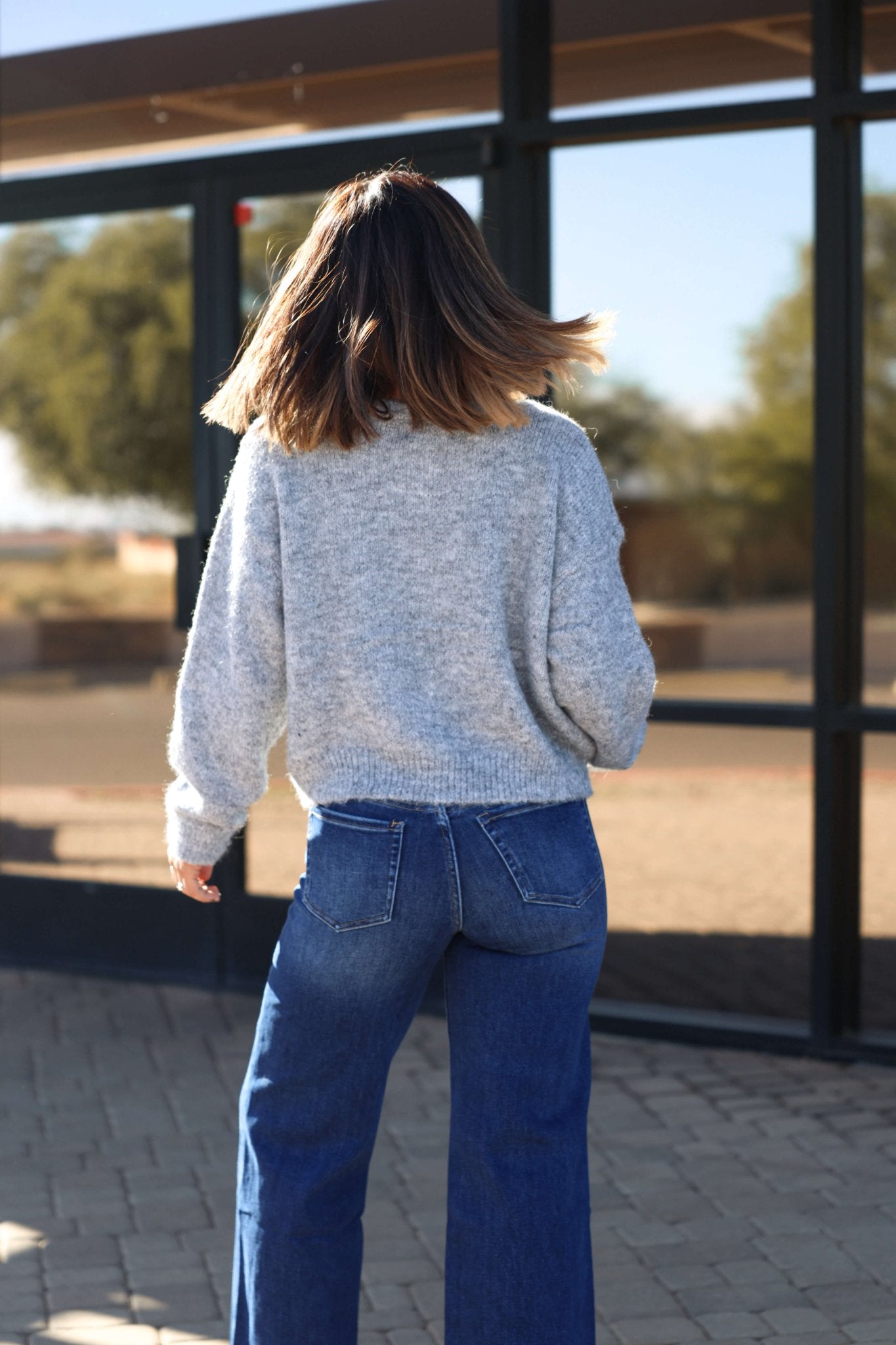 A woman wears the Lucille Grey Button Down Sweater Cardigan and blue jeans, standing outdoors with her back to the camera, turning her head.