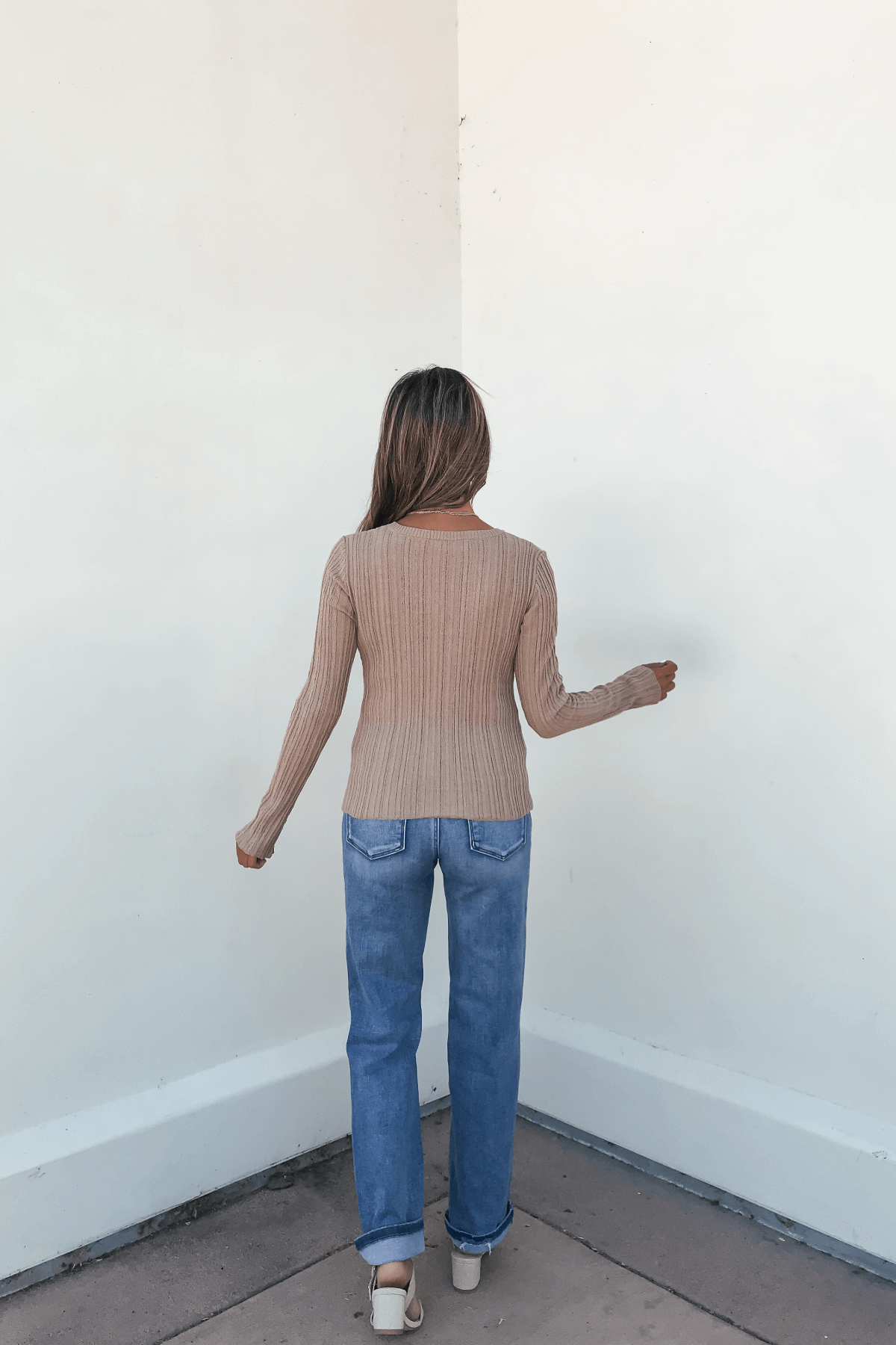 Woman with long hair wears a Margot Taupe Button Front Ribbed Top and blue jeans, facing a white wall with her back to the camera.