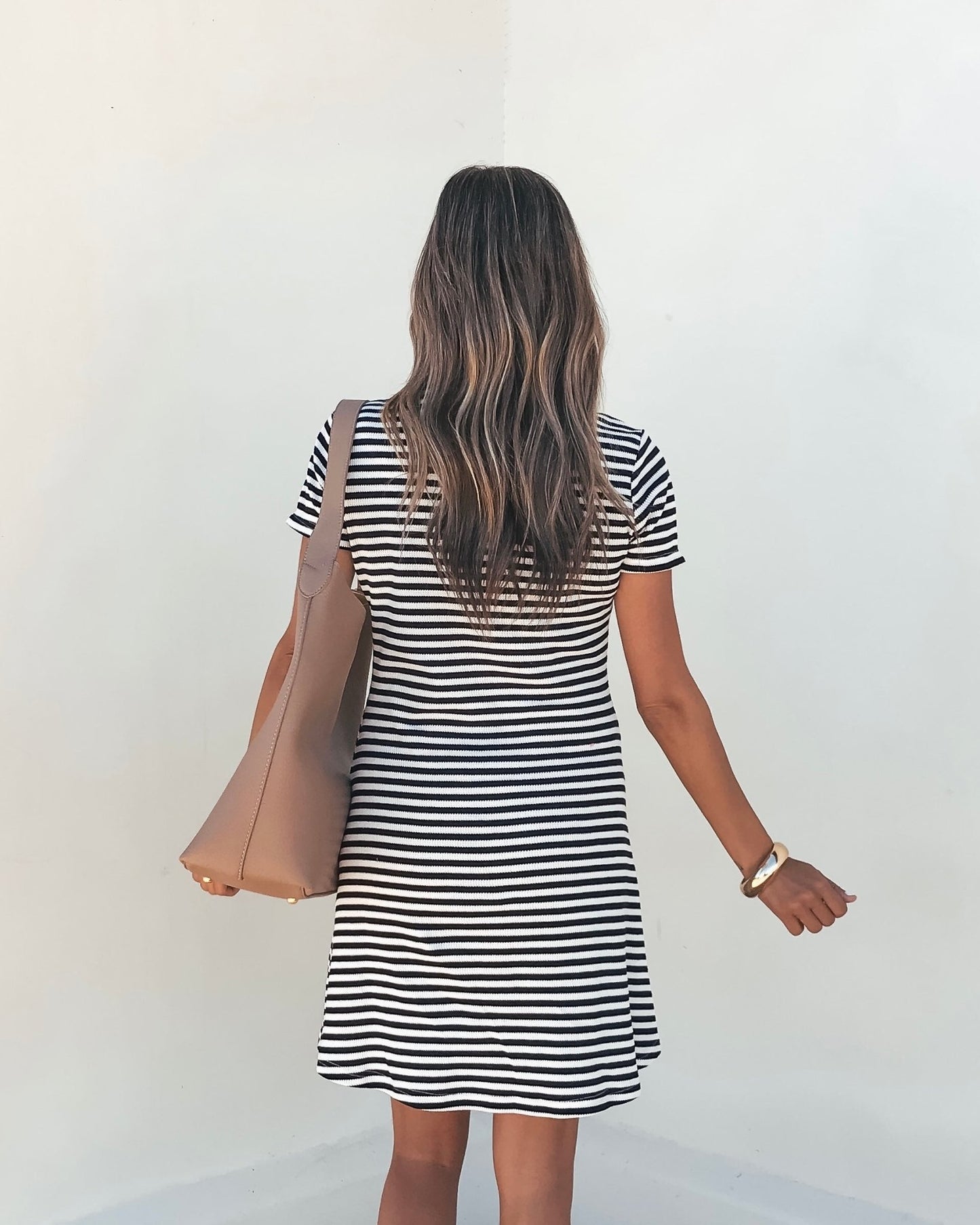 A woman with long wavy hair faces a white wall wearing the Lucy Black and White Striped Mini Dress, carrying a tan tote bag.