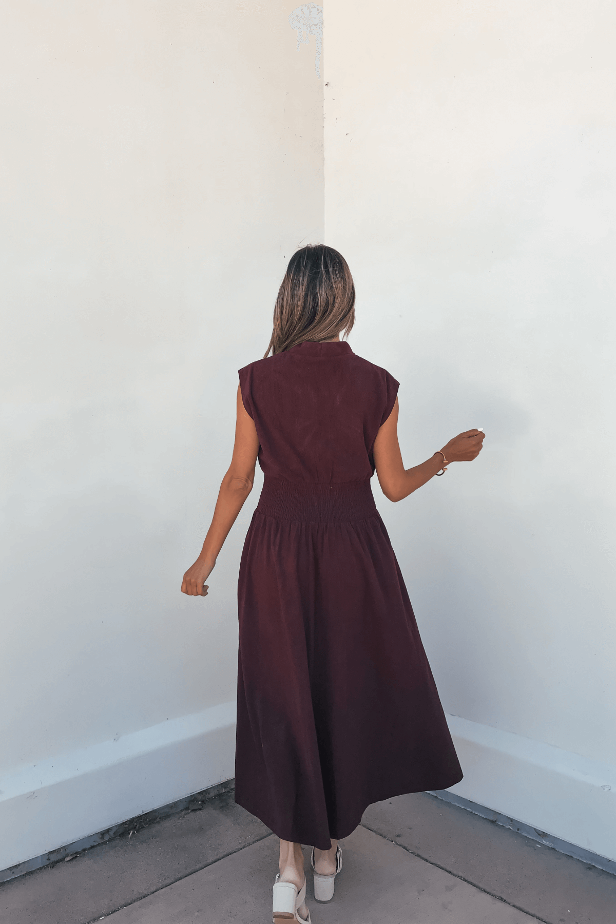 Woman in the Margot Wine Corduroy Zip Front Midi Dress stands with her back to the camera near a white wall.