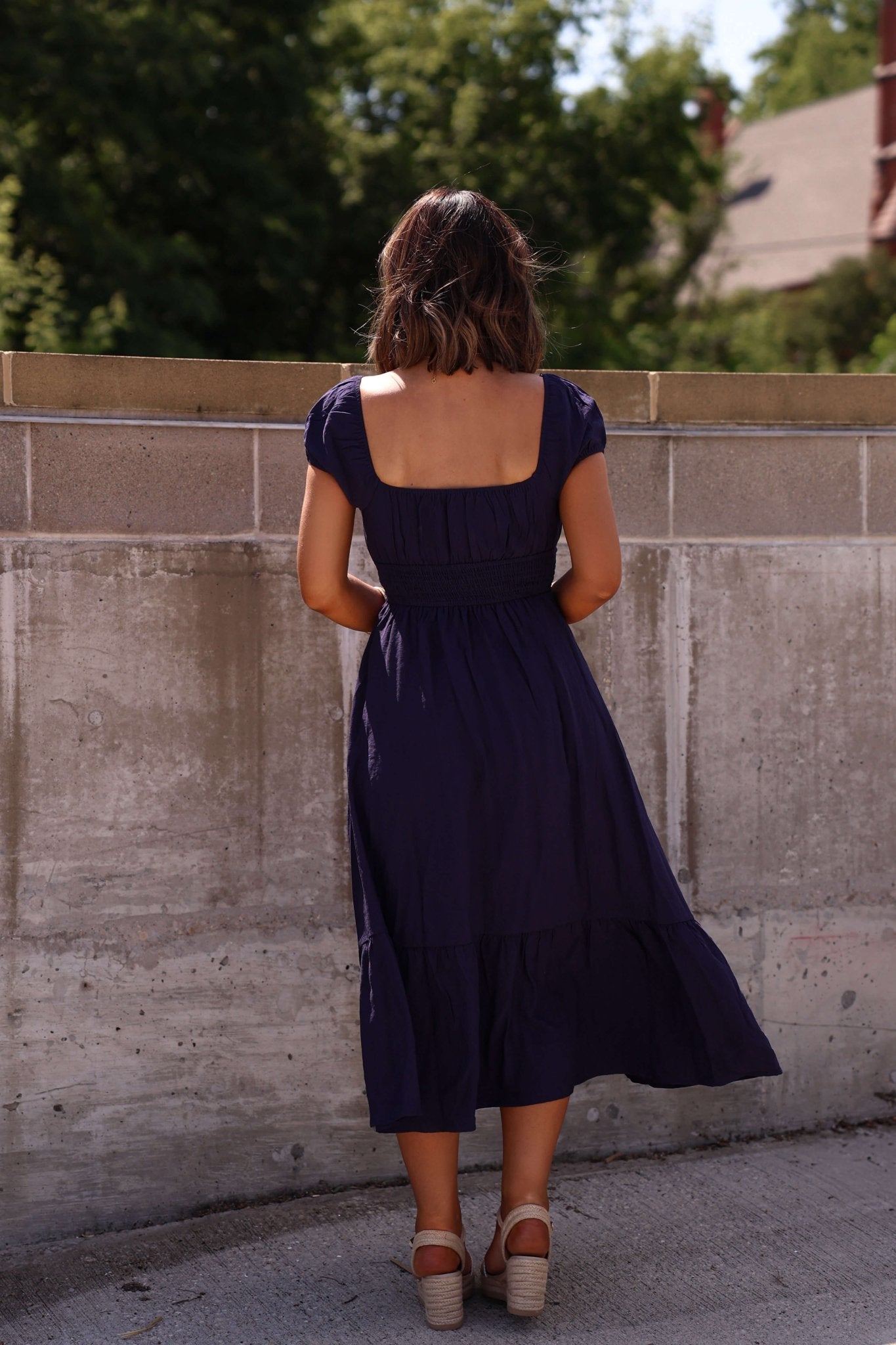 Woman wearing the Marielle Navy Tiered Midi Dress stands in sandals, facing away by a concrete wall with trees and a building behind.