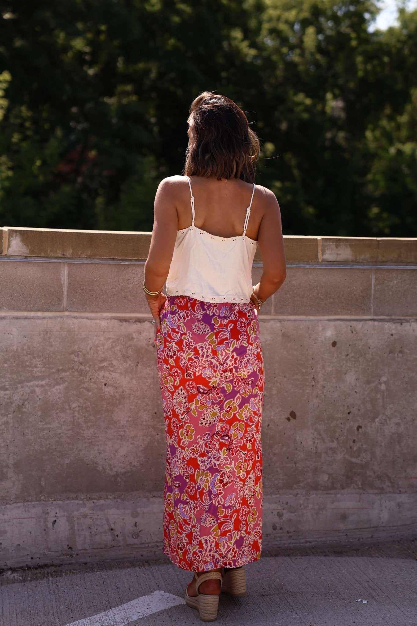A woman in a white top and the Marlowe Afternoon Pink Floral Print Midi Skirt stands outdoors, back to camera by a concrete wall.