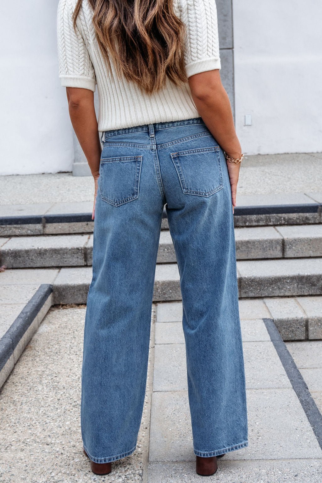 A woman in a cream sweater and Medium Wash Baggy Wide Leg Jeans stands outdoors on concrete steps, her long wavy hair flowing behind.