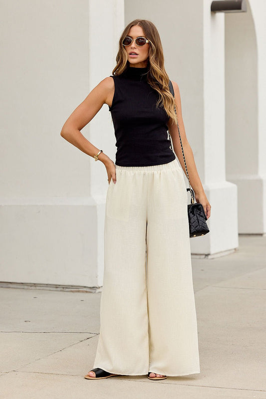 Woman in sunglasses wears the Meridian Mock Neck Tank Top - Black with wide-leg white pants, holding a small purse on the sidewalk.