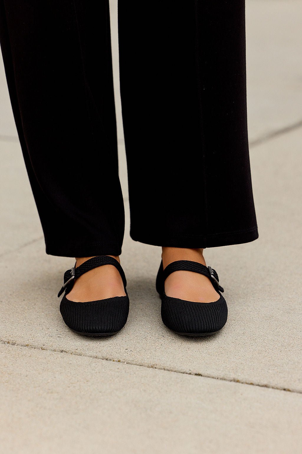 Person wearing black pants and MIA Brita Ballet Flats in Black with a Mary Jane strap, standing on a light concrete surface.