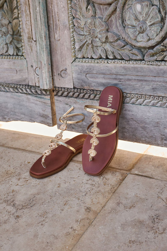 MIA Dorcie Strappy Sandals in gold with adjustable ankle straps and maroon soles, shown on stone before an ornate wooden door.