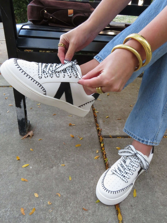 Wearing jeans, someone ties MIA Itzia Black and White Stitched Sneakers while sitting on an outdoor bench.