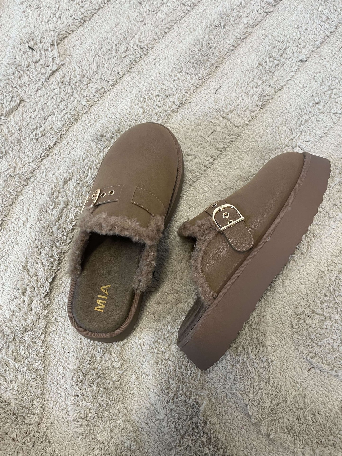 Two MIA Sugary Taupe Sherpa Mules with gold buckles rest on a textured beige rug.