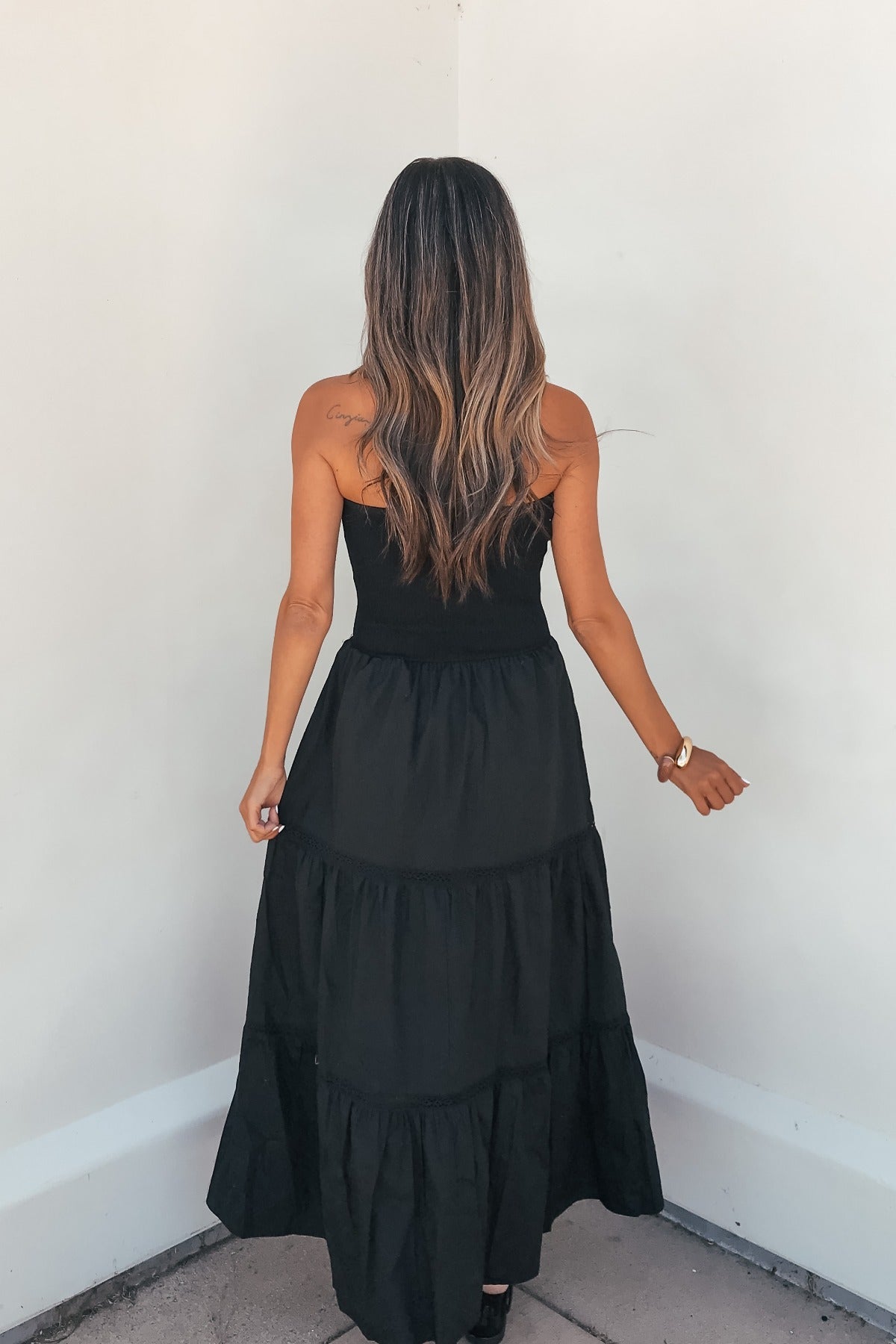A woman with long wavy hair models the Midnight Ease Strapless Tiered Maxi Dress - Black, facing a white corner from behind.