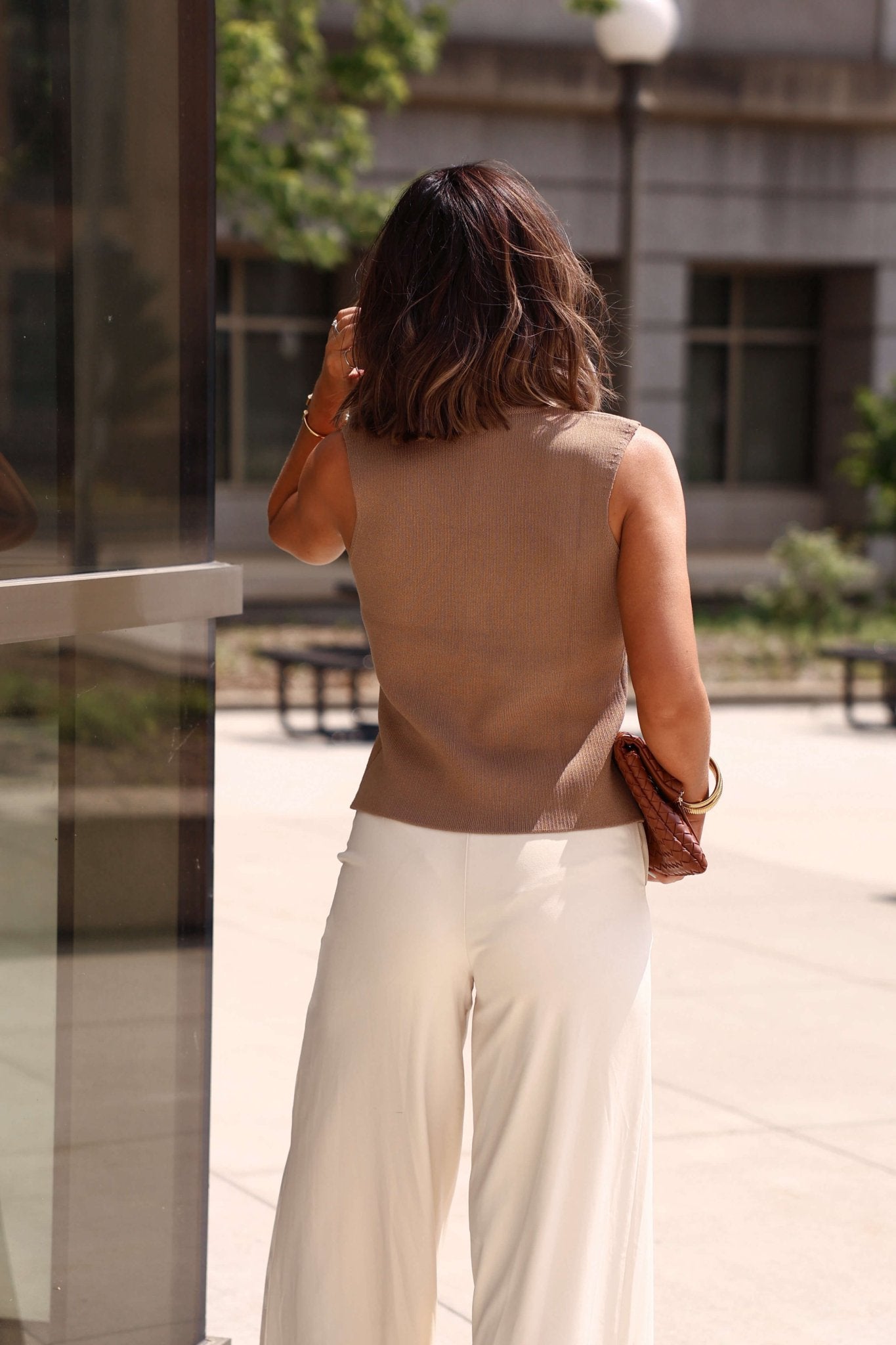 A woman with shoulder-length hair stands outdoors wearing the Mocha Button Up Sweater Vest and white pants, holding a brown clutch.
