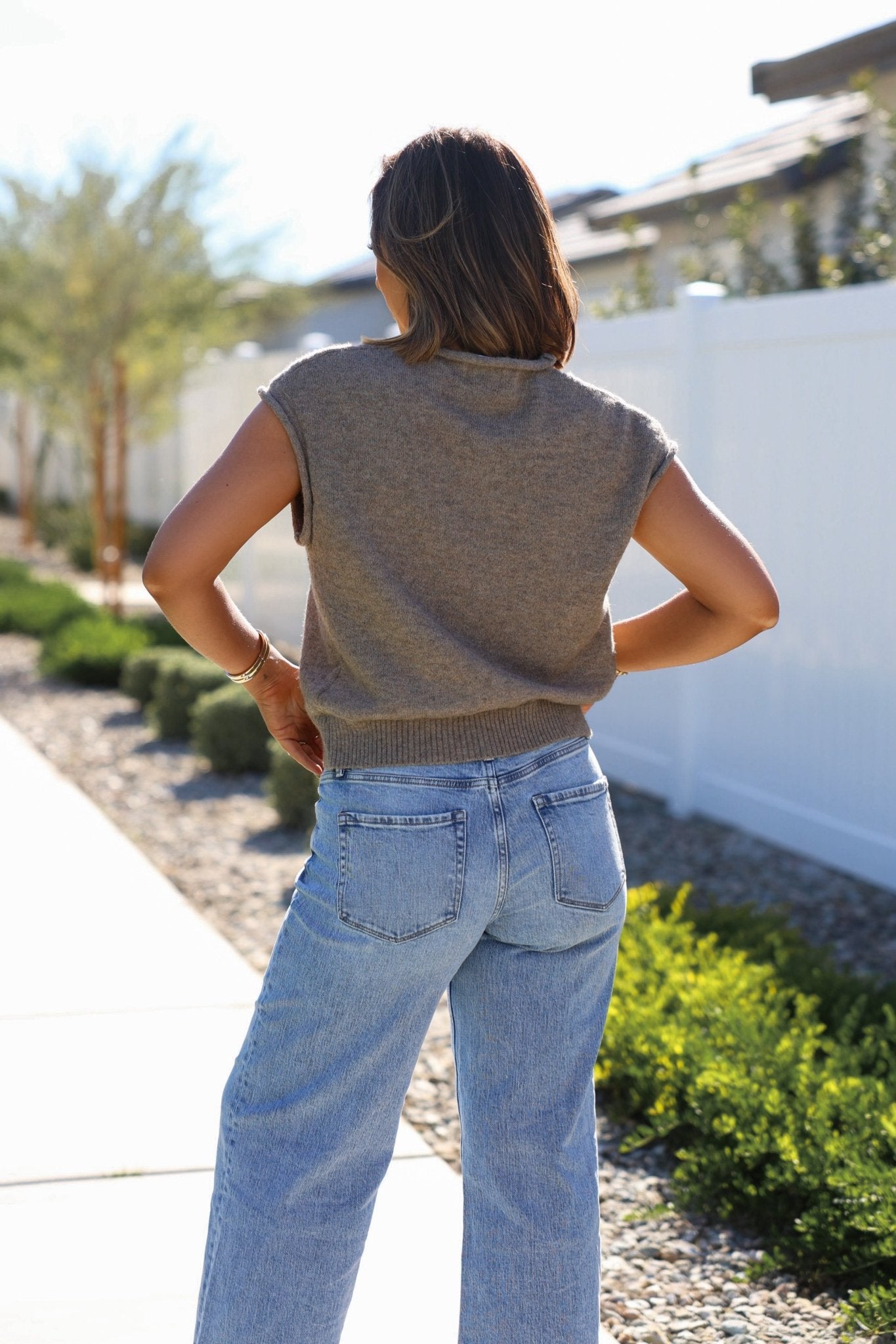A woman stands outdoors with her back to the camera, wearing a Mocha Cap Sleeve Button Down Top and light blue jeans.