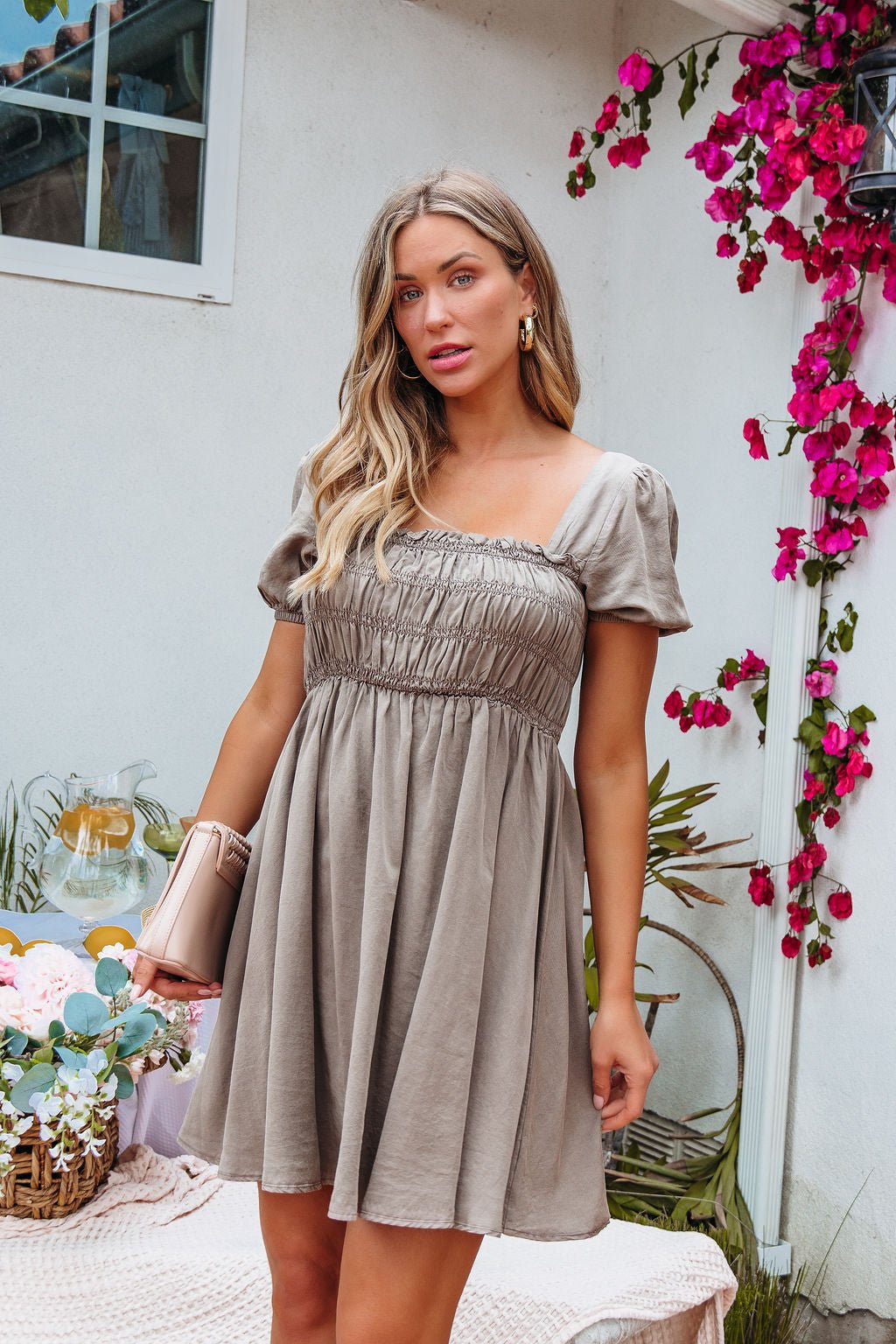 A woman in the Mocha Ruched Puff Sleeve Mini Dress stands outdoors by pink flowers, holding a clutch with a picnic behind her.